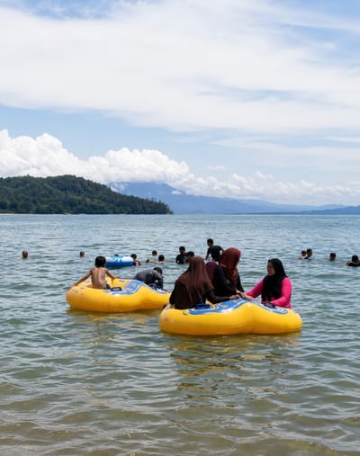 In Lumban Bul Bul, Indonesia, on a body of water, a group of people are engaged in recreational activity. Several individuals are in the water, with some using yellow inflatable tubes. In the foreground, two large yellow inflatable tubes are prominent. One tube has a young boy with dark hair sitting in it, facing away from the camera, with his arms resting on the edges. The second tube, positioned to the right, carries three women wearing hijabs. One woman in the center has a brown hijab, the woman to her left has a dark brown hijab, and the woman to her right is wearing a bright pink top and a black hijab. They appear to be interacting and are positioned side-by-side in the tube.
In the background, other people are visible in the water. Some are swimming, while others are also near inflatable tubes, including one blue tube. The water is calm with gentle ripples. The environment features a shoreline with lush green hills and trees in the distance, leading to mountains. The sky is partly cloudy with white cumulus clouds against a light blue expanse. The overall scene depicts a sunny day at a recreational water location.