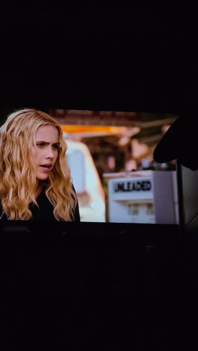 A woman with blonde wavy hair and light skin is in the foreground of the image. She is wearing a black jacket. Behind her is a white truck and an "UNLEADED" sign. An orange object is to the left of the truck. The location is not discernible from the provided image.