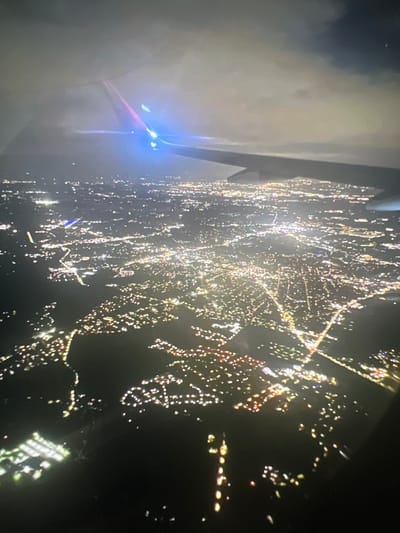 An aerial view at night shows a city illuminated by countless lights, with the wing of an airplane visible in the foreground and a portion of the tail with lights is visible in the upper portion of the image. The city lights form a dense network, suggesting a populated area. There is a notable presence of clouds that are illuminated by the city lights from below and by some external light source from above. The image depicts a wide expanse of urban development stretching into the distance, with varying densities of lights indicating different types of infrastructure and residential areas. Some parts of the city show distinct patterns of light, possibly outlining streets or larger buildings.