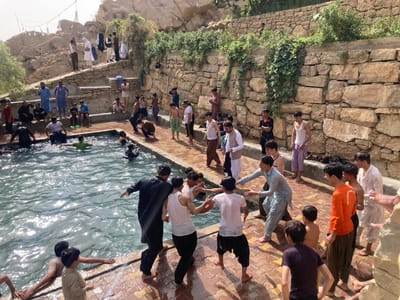 This image captures an outdoor recreational scene in Killi Malik Sher Mohammad, Pakistan, centered around a rectangular pool of water. Approximately 30-40 males, primarily youths and young adults, are present in and around the pool area. The pool, featuring clear, rippled water, is surrounded by reddish-brown brick paving. Dry-stacked stone retaining walls, partially covered with green foliage, rise in terraced levels behind the paving, leading up to a rocky, barren hillside in the background. Utility lines or decorative lights are visible strung against a bright sky in the upper left.
Several individuals are submerged in the pool, swimming or wading. On the wet brick paving bordering the pool, a cluster of males is gathered. In the mid-ground, two young males are engaged in a dynamic, playful interaction with extended arms; one wears a white tank top and dark pants, the other a light blue shalwar kameez. Other males on the paving are observing, standing in various states of dress including white tank tops, traditional shalwar kameez, and an orange t-shirt. Higher up, on the stone steps and ledges, numerous other males are seated or standing, some in shalwar kameez, others shirtless, overlooking the pool. The scene is brightly illuminated by natural sunlight, casting visible shadows.