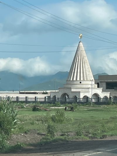 This is a medium-distance, eye-level, horizontal shot of a distinctive white building with a conical roof topped by a golden crescent, located in Rya Taza, Armenia. The building appears to be a religious or cultural site, possibly a temple or shrine, given its architectural style. The conical roof is ribbed, and the building features a series of arched openings on its lower level, enclosed by a decorative fence. Behind the building, mountains are visible, partially obscured by clouds. Power lines stretch across the bright blue sky, which is dotted with scattered clouds. The foreground consists of a grassy area with some scattered debris and vegetation. The scene is static, with no people or discernible activity.