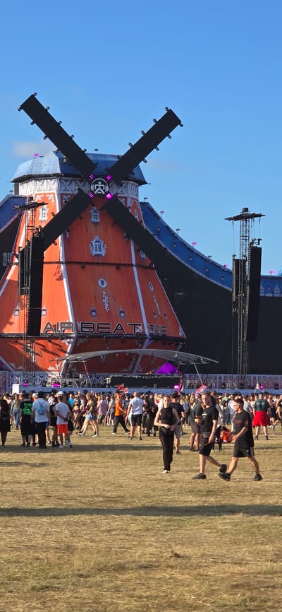 The image shows a crowd of people gathered in an outdoor field. In the background, a large, orange, windmill-shaped stage structure is visible, with the words "AIR BEAT ONE" prominently displayed on it. The windmill has black blades and is decorated with white window details. There are also large speaker towers on either side of the stage. The sky is clear blue. The people in the foreground are walking and standing, appearing to be attending an outdoor event or festival. The ground is covered in dry grass. The festival is the Airbeat One festival, which takes place annually in Neustadt-Glewe, Germany.