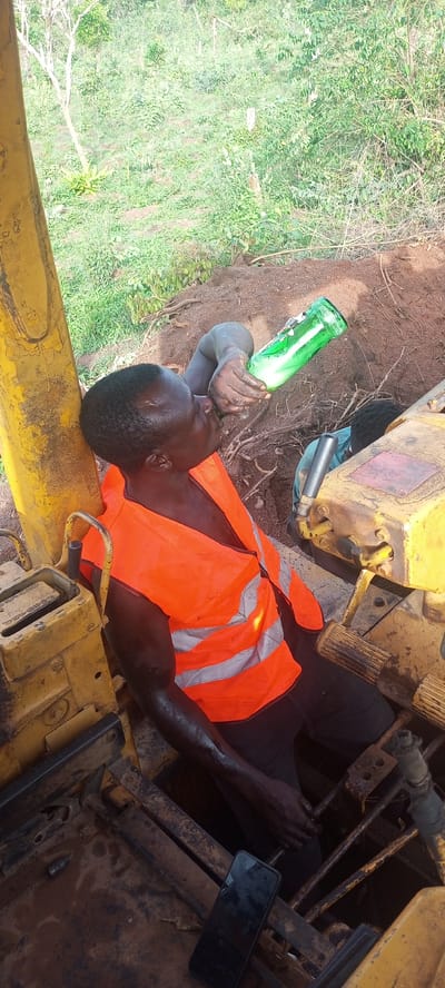 The image depicts a man taking a break while working with heavy machinery in Otunade, Nigeria. The primary subject is a dark-skinned man, seen from a high angle, seated within what appears to be the cabin or operator's area of a yellow excavator or bulldozer. He is wearing a bright orange safety vest with reflective grey stripes.

The man is actively drinking from a green glass bottle that contains a frothy, light-colored liquid, holding it to his mouth with his right hand. His skin, particularly on his arms and face, appears dirty or sweaty, indicative of physical labor. A black mobile phone with a dark screen lies on the dirty metal floor of the machinery, near his left leg.

Partially visible in the background, near the machinery's controls and within a dug-out area, is the head and shoulder of another person. The immediate surroundings consist of disturbed brown earth and exposed tree roots. Beyond the machinery and excavation, the background is filled with abundant green vegetation, including trees and dense foliage, characteristic of a natural, rural setting. The scene is brightly lit, suggesting it is daytime. No distinct text is visible on the bottle, machinery, or phone screen. The overall impression is of a worker resting during a task in an outdoor construction or excavation environment.