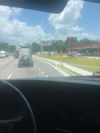 The image depicts a view from the driver's perspective within a truck, looking out at a highway scene. In the foreground, the dashboard of the truck is visible, with a digital clock displaying "10:44". The road ahead shows multiple lanes of traffic, with several trucks and cars. On the right side of the road, a Racetrac gas station is visible with its distinctive red and white canopy. A gas price sign indicates "Regular 3.54". Further down the road, a billboard advertises "BEST in OMMERCIAL PEST CONTROL YATES-ASTRO". On the median, a small fire is burning amongst some debris. The sky is blue with scattered white clouds. The scene appears to be on a highway in Savannah, Georgia, United States.