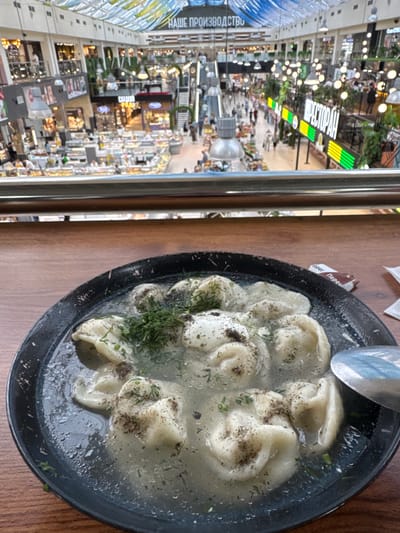A top-down shot shows a dark bowl filled with pelmeni, a type of Russian dumpling, in broth. The pelmeni are light-colored and sprinkled with black pepper and finely chopped green herbs, likely dill. The broth appears clear with some oiliness on the surface. A large, shiny metal spoon rests in the upper right portion of the bowl, partially submerged in the broth. The bowl sits on a wooden surface, possibly a table.
To the left of the bowl, a reflective surface, likely a glass window or railing, shows a blurred reflection of an indoor marketplace or shopping mall. Visible in this reflection are rows of shops, bright lights, greenery, and signs with text in Cyrillic. One sign reads "РЕСТОРАН" (RESTORAN) in prominent white and neon green lettering, indicating a restaurant area. Another sign to the left has Cyrillic text that includes "НАШЕ ПРОИЗВОДСТВО" (NASHE PROIZVODSTVO), meaning "OUR PRODUCTION." The overall scene suggests a meal being enjoyed in a food court or restaurant within a large commercial space. The lighting appears to be artificial, typical of an indoor environment, and there are no indications of time of day or weather. The focus is on the food, with the background providing context of the dining environment.