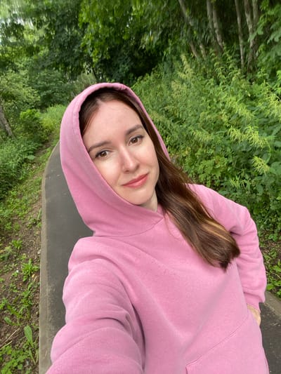 A young woman with long brown hair and light skin is taking a selfie. She is wearing a pink hooded sweatshirt and is standing on a paved path in a wooded area. The path appears to be a walking trail, and the surrounding vegetation is lush and green. The trees are tall and dense, indicating a natural setting. The overall lighting suggests it is daytime, likely overcast or early morning/late afternoon, as there are no harsh shadows. The location is identified as Melnikovo, Russia.