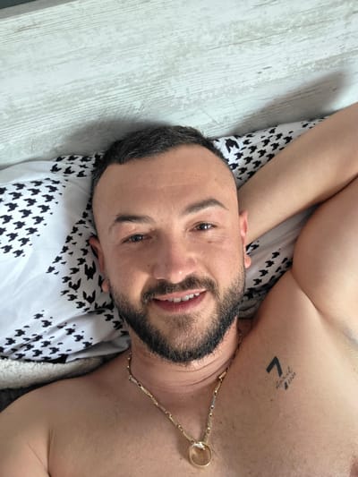 An adult male is depicted in a close-up, chest-up shot, appearing to lie in a bed in Suvorovo, Bulgaria. He has short dark hair with faded sides, a dark beard, and a mustache. He is smiling faintly, making direct eye contact with the camera. His bare upper torso and shoulders are visible, and he wears a gold-colored chain necklace with a ring-like pendant. A tattoo on his left shoulder displays a stylized '7' above 'April 2020'. He is resting on a pillow with a black and white repeating pattern resembling bird or houndstooth shapes. A white wooden-paneled surface is visible behind his head, and a portion of a light-colored, possibly sherpa-lined blanket is seen near his left side.