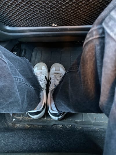 The image, captured from a high-angle perspective, showcases the lower legs and feet of an individual situated inside a vehicle. The person is wearing dark, possibly black or charcoal grey, denim jeans with raw or frayed hems. Their feet are clad in white sneakers featuring distinct black stripes on the sides, characteristic of Adidas footwear. The visible surface where the feet rest is a dark grey or black rubber floor mat with a raised grid pattern. To the left, a section of the car's interior reveals a black mesh-textured mat. To the right of the person's feet, a dark, possibly carpeted area of the car floor is visible, separated by a dark, rigid interior trim or structural element. This carpeted section appears somewhat dusty or lightly soiled. The lighting is bright and diffused, indicating daytime conditions. There is no specific activity depicted other than the person occupying the space within the car. No discernible text beyond the faint Adidas branding on the sneakers is apparent. The scene provides a commonplace glimpse into a car's interior in Ans, Belgium.