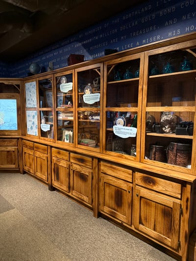 A medium shot depicts a large wooden display case, likely located in a museum or exhibition in Powell, United States of America, given the information plaques and displayed objects. The case is filled with various items, including blue glass orbs, a jar of what appear to be rocks, and several books. Additionally, information plaques about polar bears are visible, suggesting a natural history or wildlife-themed exhibit. The wall to the left has dark blue text on a lighter blue background, and the wall to the right is carpeted, which is likely one of the building’s structural supports.
