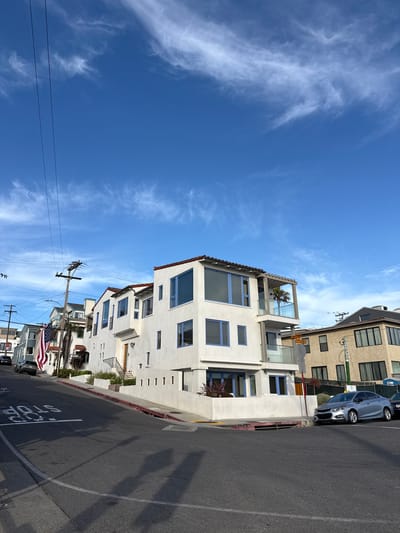 This is a medium-distance, eye-level, outdoor, full shot of a residential street corner in Hermosa Beach, California, USA. The sky is a clear, bright blue with wispy white clouds. A power pole with overhead wires is visible on the left side of the frame. The street is paved, with a white stop line painted on it in the foreground.
The primary subject is a modern, white, three-story house with blue-trimmed windows and a flat roof. An American flag is displayed on a pole to the left of the house. Adjacent to the white house is a tan, three-story building. A silver sedan is parked on the street in front of the tan building. Another dark-colored vehicle is parked further down the street on the left. Street signs are visible on a pole near the intersection. The overall scene depicts a sunny day on a residential street with distinct architecture.