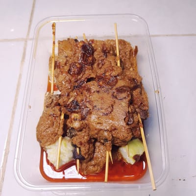 The image displays a ready-to-eat meal of Indonesian satay skewers contained within a clear, rectangular plastic take-out box. The skewers, made of bamboo, hold pieces of dark meat or organ meat, which are generously smothered in a thick, savory brown sauce. This sauce, likely a peanut-based variant, has a rich texture and is topped with crispy, dark brown elements, strongly resembling fried shallots (bawang goreng).

Accompanying the satay are several pieces of white, compressed rice cakes, commonly known as lontong or ketupat in Indonesian cuisine, providing a starchy complement to the meal. A distinct section of the container holds a bright red chili sauce, offering a spicy contrast to the savory main dish. The sauces and rice are somewhat mixed, indicating they are packaged together.

The clear plastic container, typical for food delivery or take-away, shows some condensation and reflections from artificial light sources. It rests on a simple white surface, possibly a tiled countertop or floor, identifiable by the subtle grout lines. The overall presentation suggests a prepared meal ready for consumption. No individuals or written text are visible in the photograph, and the time of day is not discernible. The dish is consistent with Indonesian culinary traditions found in locations like Lampeudaya.