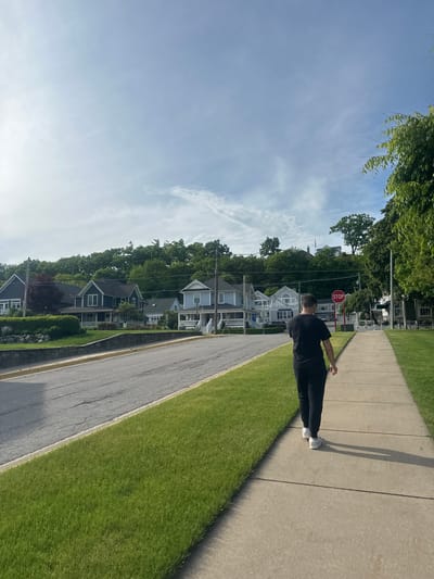 In Harbor Springs, United States of America, a person wearing a black t-shirt and black pants walks on a concrete sidewalk alongside a manicured lawn and a road. The person is walking away from the camera, casting a shadow on the sidewalk. Behind the sidewalk, a line of houses with varying architectural styles is visible, nestled against a lush green hillside covered with trees. The sky is a pale blue with scattered white clouds. A red stop sign is visible on the right side of the road, near the intersection. The scene depicts a suburban or residential area with a clear sky and well-maintained surroundings.