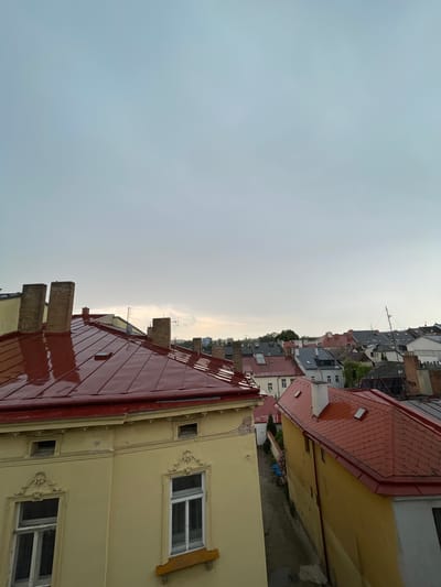 The image shows a view of rooftops in Jihlava, Czechia, likely from an elevated position such as a balcony or another building. In the foreground, a large building with a red, glossy metal roof and yellow facade is prominent. This building has multiple brick chimneys and several windows, some with decorative molding. Adjacent to it is another building with a red tiled roof. In the background, a cluster of various rooftops can be seen, indicating a densely populated urban area. The sky is overcast with a light gray hue. The overall scene suggests a residential or mixed-use district within the city.