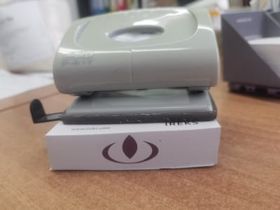 The image shows a close-up of a light gray hole puncher placed on top of a stack of white business cards. The hole puncher has "SXD P-817" embossed on its side. The business cards have "www.ireks.com" printed on one side, along with the IREKS logo and the word "TREKS". The scene is set on a wooden desk. No people are visible. The image does not provide any information about the city of Dobrodan, Bulgaria.