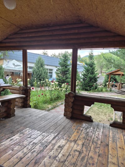 The image captures a view from a wooden porch, looking out onto a garden area with a building in the background. The porch itself is constructed from logs, with a wooden floor that appears weathered. There is a wooden bench on the right side of the porch.

Beyond the porch, a garden is visible, featuring green trees and a variety of flowering plants, including roses with red and yellow blooms. A paved walkway with a red and white pattern runs through the garden. In the background, a light-colored building with a blue roof is partially visible. A white tent with a black bird logo is set up in front of one of the buildings.

A person is visible near the tent, wearing dark clothing. Another person is sitting on a wooden structure in the garden, further away.

The lighting suggests it is daytime, likely with overcast skies as there are no strong shadows. The scene appears to be a residential or recreational area, possibly a guesthouse or a private garden. The overall impression is one of a rustic and natural setting.

Visible text includes Russian Cyrillic characters on the tent.