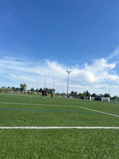 The image depicts a sunny day at a soccer field in Champigny-sur-Marne, France. A group of individuals are present on the field, seemingly engaged in practice. The environment includes the soccer field, marked with white and blue lines, stadium lights, and surrounding buildings. The sky is clear with some scattered clouds. Two jet trails are visible.