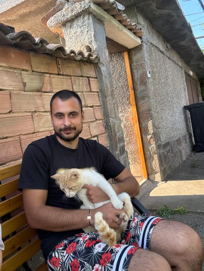 In Kozlovets, Bulgaria, a man with dark hair and a beard, wearing a black t-shirt and floral shorts, sits on a wooden bench outdoors. He is holding a light-colored cat with orange markings, which is nestled in his arms. The background features a brick wall and a stone building with a corrugated roof. Sunlight casts shadows on the ground and building.