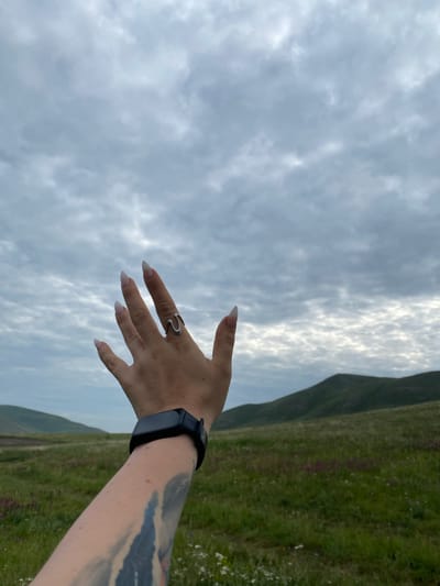 The image shows a person's hand and forearm reaching out towards a cloudy sky. The hand has long, pointed, light-colored fingernails and is adorned with a silver ring shaped like a wave. A black fitness tracker is visible on the forearm, which also features a tattoo. The background depicts a rolling, grassy landscape with distant green hills under a heavily clouded sky. The location is identified as Abzanovo, Russia.