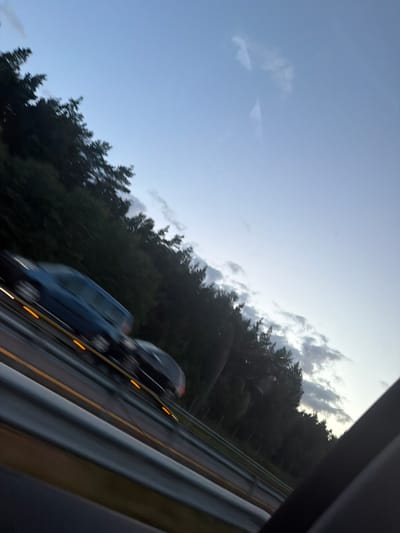 The image depicts a highway scene at dusk or dawn, with a clear sky and scattered clouds. A dark-colored car is in the foreground, moving on the highway. The car is partially obscured by motion blur, but its headlights and taillights are visible as streaks of light. To the left of the highway, a dense line of dark green trees forms a natural barrier. The highway appears to be a multi-lane road with guardrails and yellow lane markers. The overall impression is that of a vehicle traveling at speed on a rural or semi-rural highway in Berg i Østfold, Norway, during twilight hours.