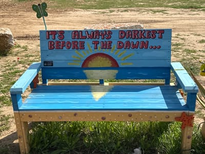 A wooden bench, painted blue and tan to look like an ocean scene, sits in the grass outside of the community garden in Re'im, Israel.  The bench is decorated with a painted sunset and the words, "It's always darkest before the dawn..." in red, yellow, and black. A green butterfly with a blue stick is protruding from the top of the bench back. There is a red starfish on the right side of the bench, and the grass surrounding the bench is mostly green with some brown patches. The ground around the bench is made of dirt and sand. The location is a public park space, the coordinates are 31.398 latitude and 34.47 longitude.