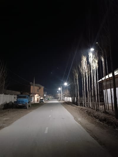 A nighttime street scene features a long road illuminated by evenly spaced streetlights. A blue truck is parked on the left side of the road, near a fence and a building. On the right, tall, thin trees line the edge of the road next to a house with a white wall. Several cars are parked farther down the street, adding to the quiet, residential atmosphere.
