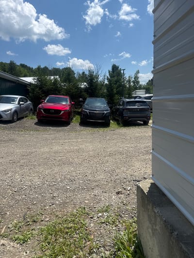A daytime exterior shot captures a gravel parking lot with three cars parked in front of a dense line of evergreen trees. To the left, a silver sedan is partially visible. In the center, a red SUV is parked. To the right, a dark-colored SUV is parked. A white, corrugated metal building runs along the right edge of the frame, obscuring part of the scene. The sky is blue with scattered white clouds. The scene appears to be located in the Village of Johnson City, United States of America, possibly a commercial or industrial area, given the presence of the building and the arrangement of vehicles.