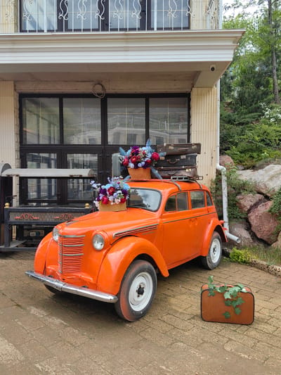 The image depicts an orange Moskvich 401 parked on a brick-paved area in front of a two-story building. The car is adorned with two baskets of artificial flowers, one on the hood and another on the roof, which also holds a stack of three vintage suitcases. A brown suitcase with green vines on top sits beside the car. The building behind the car features a cream-colored facade with a large black-framed window and a decorative balcony. Lush greenery, trees, and a rocky landscape are visible in the background. The location is Luchistoe, Ukraine.