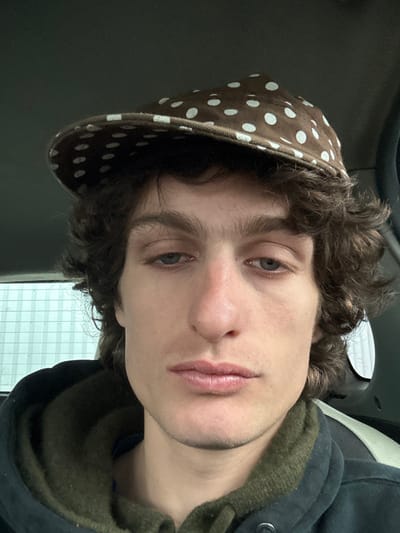 A close-up, frontal portrait of a male individual is captured within what appears to be the interior of a vehicle, situated in Vitry-sur-Seine, France. The subject, possessing medium-toned skin, dark wavy hair, and blue eyes, maintains a neutral facial expression while looking directly towards the camera. He wears a brown flat cap adorned with white polka dots, a dark-colored hooded outer jacket, and an olive-green hooded garment beneath. The background on the left reveals a window through which a light-colored structure with a prominent grid or horizontal line pattern is visible. To the right, a dark, indistinct vehicle interior component is partially discernible. The lighting is even, suggesting an enclosed space with diffused external light.