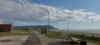 This media file depicts a scenic coastal path with a beach and ocean in the background. In the foreground, a paved walkway lined with lampposts curves away from the viewer, leading towards a green, hilly coastline. To the left of the path, there is a modern, wooden structure with an elevated platform and stairs. A white, slatted bench is positioned on the right side of the walkway, overlooking the beach. A wooden trash receptacle is next to the bench.
The beach is wide and sandy, with gentle waves lapping at the shore. In the distance, several rock formations rise from the ocean. The sky is filled with a mix of white, fluffy clouds and patches of blue, suggesting a partly cloudy day. The sun appears to be moderately high in the sky, indicating it is likely daytime, possibly late morning or early afternoon.
The setting is a well-maintained public space, possibly a park or promenade, designed for recreation and enjoying the coastal view. The overall mood is tranquil and serene, inviting relaxation and contemplation of the natural beauty. There is no visible text in the media file.
