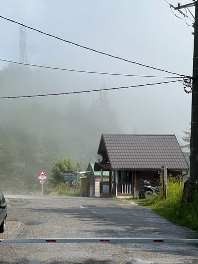 The image depicts a foggy mountain road scene in Trešnjevik, Montenegro. In the foreground, a white barrier is lowered across a gravel road, suggesting a checkpoint or entrance. To the right of the road, a wooden house with a dark brown metal roof is situated, and in front of it, a black ATV is parked. A smaller wooden structure with a green roof is to the left of the main house. Several road signs are visible: a triangular sign with a red border and a black depiction of a wild animal, indicating a wildlife crossing, and a circular sign with a red border and the number "30" within, signifying a speed limit. A larger blue sign indicates "HOTEL KOMOVI ANDRIJEVICA" with additional text and a phone number. Further back, the road curves and disappears into dense fog that envelops a lush forest. A tall, lattice-like transmission tower is faintly visible through the mist in the distance, and power lines crisscross the sky. The overall atmosphere is damp and overcast, with wet patches on the road surface and the vegetation appearing vibrant green.