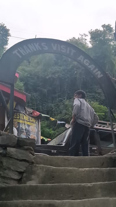 In Malana, India, a person stands on concrete stairs looking towards a dark archway with the words "THANKS VISIT AGAIN" inscribed on it. The archway is suspended over a narrow path. To the left of the archway, a building with red and yellow awnings is visible. A sign on the building reads "STONE CAFÉ" and advertises "BREAKFAST - LUNCH - DINNER," with images of "MOMOS" and "NOODLES." Prayer flags in various colors are strung between the building and trees. Behind the person, a dark-colored vehicle is parked. The background is dominated by lush green foliage covering a hillside. The sky is overcast.