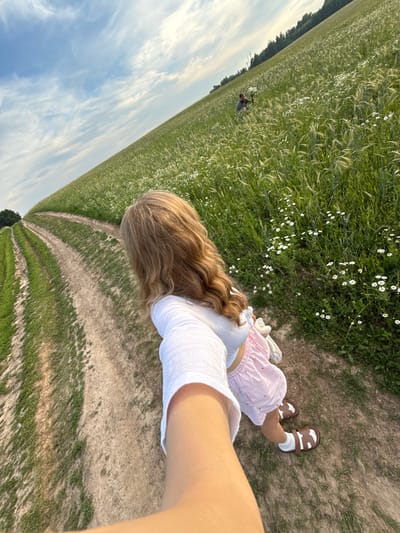 A person with long, wavy blonde hair is taking a selfie while walking on a dirt path through a field of green grass and white wildflowers. The person is wearing a white crop top and a pink and white striped skirt, with white socks and brown sandals. Another person is visible in the distance in the field, appearing to be picking flowers. The scene is set in a rural area with a gently sloping hill and a line of trees in the background. The sky is blue with scattered white clouds. The location is identified as Сновицы, Russia.