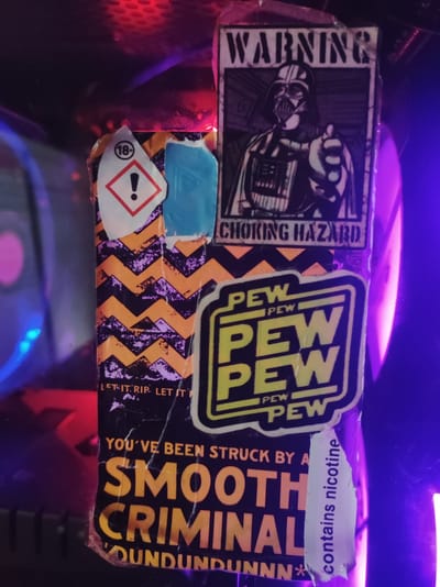The image shows a close-up of a vape juice bottle with various stickers. One sticker features Darth Vader with the text "WARNING" and "CHOKING HAZARD". Another sticker is a warning symbol with "18+" inside a circle and an exclamation mark in a red triangle. There are also several "PEW PEW" stickers. The bottle itself has a black background with orange and purple zig-zag patterns and text that reads "SMOOTH CRIMINAL" and "DUNDUNDUNNN*". A small white sticker on the side says "contains nicotine". The bottle is illuminated by purple and red lights, possibly from a computer case. There is no indication of the location being Roranwala, Pakistan, or any other specific city.