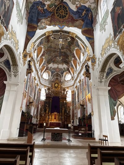 The image shows the interior of a church. The ceiling is highly decorated with paintings and gold trim. A clock and depictions of figures are visible on the ceiling. A crucifix hangs from the ceiling. Windows provide natural light. In the foreground, there is a wooden pew. In the background, there is an altar covered with a purple cloth, surrounded by ornate decorations. The location is Biberach an der Riß, Germany.