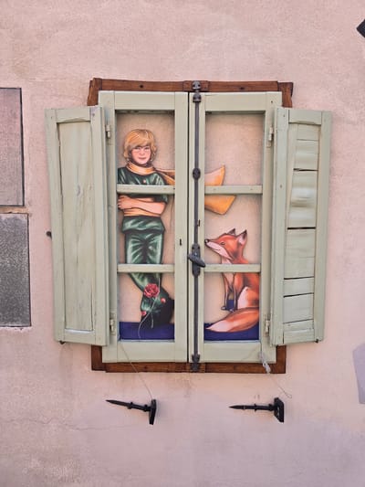 This image shows a whimsical mural painted on the exterior wall of a building in Viterbo, Italy, depicting characters from "The Little Prince." The artwork is framed by an open, pale green wooden window with shutters.
On the left side of the window, a stylized representation of the Little Prince stands with his arms crossed. He has blonde hair, a light brown scarf, and is wearing a green jumpsuit. A red rose with a long stem is painted at his feet.
On the right side of the window, a fox is depicted sitting with its head turned to the left, looking towards the Little Prince. The fox has reddish-brown fur and a bushy tail. Above the fox, two orange shapes, resembling the boa constrictor digesting an elephant from the book, are visible.
The window frame itself is rustic, with visible wood grain and signs of age. A metal latch and hinges are present on the window. Below the window, two small, black, spike-like fixtures are attached to the wall, each connected by a thin wire to the window frame. The wall itself is textured and painted in a pale pinkish-beige hue. To the left of the window, a section of a different, rectangular window with a frosted appearance is visible. The overall scene is a creative use of a window to present a painted narrative.