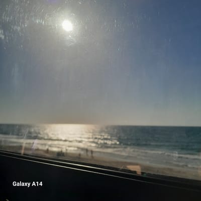 The image appears to be taken through a window. Outside, there is a view of the sky and what seems to be a body of water, possibly the sea or a large lake. The sun is visible in the sky, creating a glare. The location is indicated as Al-Mawasi, Unknown. The image was taken with a Galaxy A14 phone. There are no people visible in the image. The focus seems to be on the outdoor scenery.
