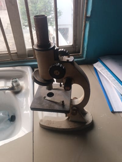 An old microscope stands on a tiled surface, near a sink and a window. The microscope is weathered, with its metal components showing signs of rust. Its beige body has visible wear. A blue file folder is to the right. The environment appears to be a laboratory or a similar setting.