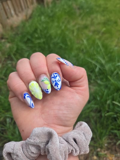 A hand adorned with intricately designed fingernails is held up against a background of blurred green grass and a wooden fence. The nails feature a Mediterranean-inspired pattern, with several nails painted white and decorated with blue floral motifs, resembling traditional ceramic tiles. Two nails prominently display detailed designs: one with a bright yellow, sculpted lemon slice, and another with delicate green leaves and smaller yellow lemon shapes on a translucent base. The thumb nail is partially visible, with a white tip decorated with blue dots. The hand is wearing a beige scrunchie around the wrist. The image appears to have been taken outdoors, in Vale of White Horse, United Kingdom, judging by the natural lighting and grassy environment.