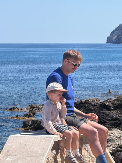 A medium shot depicts a man and a child seated together near the sea. The man, wearing a blue shirt and sunglasses, is positioned behind the child. The child is wearing a light-colored shirt, a baseball cap, and striped shorts. They are seated next to a low concrete wall. Behind them is a rocky outcropping and the sea. The location is likely in Cala Rajada, Spain, based on the coastal scenery.