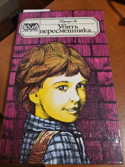 The image shows the cover of a book titled "Убить пересмешника..." which translates to "To Kill a Mockingbird...". The cover features a yellow-toned illustration of a child's face, with brown hair and a checkered shirt. The background is a pink brick wall pattern. To the left of the title, there is a label with three globes and the text "ДВА МИРА – ДВЕ СУДЬБЫ" which means "Two Worlds - Two Fates". The book is placed on a wooden surface, likely a table. The image does not contain any information about the city of Anadyr, Russia, nor does it depict any forensic details or actions.