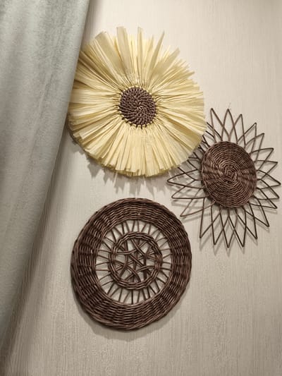The image shows three decorative wall hangings on a textured, light-colored wall. The wall hangings are circular. The largest, positioned at the top left, is made of light yellow paper strips radiating outwards from a brown woven center, resembling a stylized sun or flower. To its right, a smaller, darker brown woven hanging also resembles a sun with pointed rays. Below and to the left of this, a third woven circular piece features a more intricate, basket-like pattern. A grey, soft fabric, possibly a curtain or upholstery, is draped on the left side of the image, obscuring a portion of the wall and the leftmost hanging. The lighting suggests an indoor setting, casting subtle shadows from the wall hangings onto the textured surface. There are no people or animals present in the image. There is no information available to determine the location within the city of Речицы, Russia.