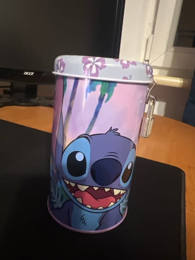 A Stitch-themed tin can with a padlock sits on a desk next to a computer monitor. The can is decorated with Stitch's face and a tropical background. The monitor is black with an "acer" logo on the front. The desk surface is a light brown wood grain. The image appears to be taken indoors, likely in a home office or bedroom. There is no indication of the location being in Skawina, Poland.