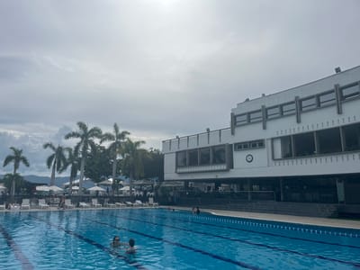 The image displays an outdoor swimming pool complex located in Floridablanca, Colombia, observed under an overcast sky with bright light in the upper right quadrant. The foreground features a large, rectangular swimming pool with clear blue water and visible dark blue lane lines. Two individuals are present in the pool: one adult, identifiable by sunglasses, and one younger individual, both partially submerged.
On the left side of the frame, behind the pool, stand multiple tall palm trees, with distant mountains visible on the horizon beyond them. An adjacent paved area by the pool contains numerous unoccupied white lounge chairs and several opened white sun umbrellas. A few indistinct human figures are visible near the lounge chairs and pool edge on the left.
The right side of the image is dominated by a multi-story, white, modern building. The structure features large rectangular windows, distinct horizontal architectural elements on its upper level, and a section with vertical slats or fins on a lower section. A circular clock with a white face and black hands is mounted on an exterior wall of the building. The building's base transitions into a darker, stepped wall descending towards the pool. Green foliage is visible around the building and behind the pool area, indicating a lush environment.