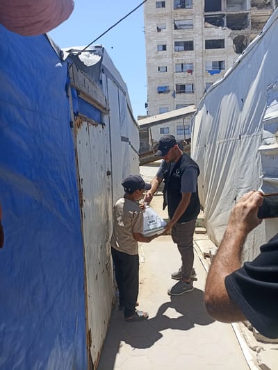 In a scene of apparent humanitarian aid distribution, a man in a dark vest over a grey t-shirt and dark pants is handing a package to a young boy wearing a tan shirt and dark pants, with a black baseball cap. The package is white and transparent, with text visible on it. The man is wearing a dark baseball cap and athletic shoes. The boy is also wearing a black baseball cap and sandals. The setting appears to be a narrow, open-air space between makeshift structures covered with blue and white tarps. In the background, a damaged, multi-story building with broken windows and visible destruction suggests a context of conflict or disaster. The sky is clear and blue, indicating a daytime scene with fair weather. The overall atmosphere suggests a moment of support and relief amidst hardship. The text on the package reads "GAZA HOT MEALS JULY 2023".