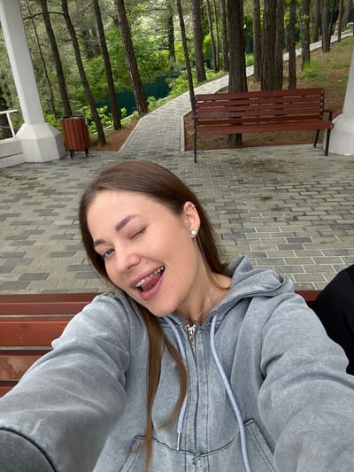 A young woman is sitting on a wooden bench, possibly in a park in Sosnovy, Russia. She is wearing a grey hoodie and has brown hair. The woman is sticking her tongue out and winking at the camera. The ground is paved with grey brick, and there are trees and another bench in the background. A trash can is visible near the trees.