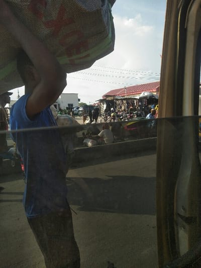 The image depicts a street scene in Awjaw, Nigeria, viewed from inside a vehicle. In the immediate foreground, a person, likely male, wearing a blue t-shirt and dark lower garment, is seen from the back and left profile, carrying a large, light-colored woven sack with red lettering on their head, supported by their raised right arm. A smaller white bag is slung over their left shoulder. The right side of the frame is occupied by a vertical, metallic, possibly bronze-colored structural component of the vehicle.
The midground features an asphalt road with a concrete curb separating it from an elevated pedestrian or market area. Numerous individuals are present: some are walking, others are seated on the curb, appearing to engage in market transactions with various bags and goods around them. Multiple motorcycles are parked or moving within this area. Behind the people, a row of market stalls or small shops is visible, some with reddish-brown corrugated metal roofs and others with darker tarpaulin coverings. Clothing items are displayed for sale.
In the background, a white, block-style building is discernible. The sky is partly cloudy, and overhead utility lines traverse the scene. The overall environment suggests a busy, open-air market setting during daylight hours.