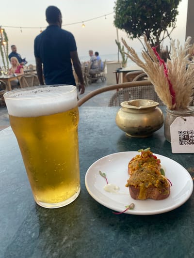 In Taghazout, Morocco, at an outdoor dining establishment near the ocean, a glass of golden-hued beer with a frothy head sits on a dark green speckled table. Adjacent to the beer is a white plate containing two pieces of toasted bread topped with a savory mixture and garnished with microgreens and a dollop of white sauce. In the background, a person in a navy blue shirt is walking away from the camera. Further back, other patrons are seated at tables, some dining and others conversing, with glimpses of the ocean and lush greenery. A decorative arrangement of dried grasses and a brass ashtray are also visible on the table. The overall scene suggests a relaxed evening at a seaside restaurant.