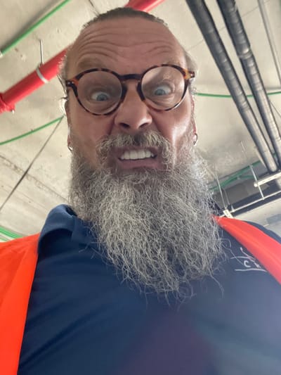 The image shows a close-up, selfie-style shot of a man with a long, gray beard and glasses. He is wearing an orange safety vest over a navy blue shirt. His expression is contorted, with wide eyes and his teeth bared in a grimace or grimace. The background appears to be an industrial or construction setting, with pipes, conduits, and concrete ceiling visible. The lighting is somewhat dim. There is no indication of the specific location within Ramot HaShavim, Israel, other than the general context of an indoor industrial area.