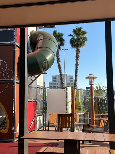 This image depicts an outdoor dining area with a playground structure visible in the background. In the foreground, there are several wooden tables and chairs, some with sunlight casting shadows on them. To the left, part of a red and brown playground structure with a green tube slide is visible. Beyond the playground, two palm trees stand tall against a clear blue sky. Further in the background, several modern buildings can be seen, indicating an urban setting. The scene appears to be taken from inside an establishment, looking out onto an outdoor patio and playground area. The location is identified as Sedaví, Spain. There are no people visible in the image.