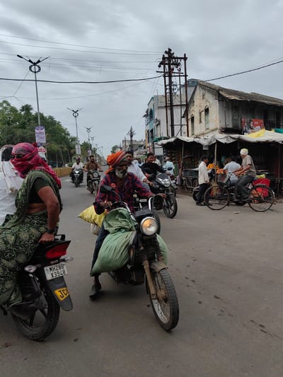 The image depicts a street scene in Malkapur, India, likely on a cloudy day. In the foreground, a person wearing a green sari is seated on a motorcycle, with a red head covering partially obscuring their face. Another person, an older man with a beard and an orange turban, is riding a motorcycle in the right foreground, with a large green sack strapped to the front of his bike. Several other motorcycles and a bicycle are visible on the street, with people riding them. On the right side of the street, there are shops with awnings, selling what appear to be fruits and vegetables. Buildings with traditional Indian architecture line the street, some showing signs of age. Electrical poles and overhead wires are prominent features of the urban environment. The road surface is asphalt, with some scattered debris. The overall impression is one of everyday life and commerce in a bustling Indian town.